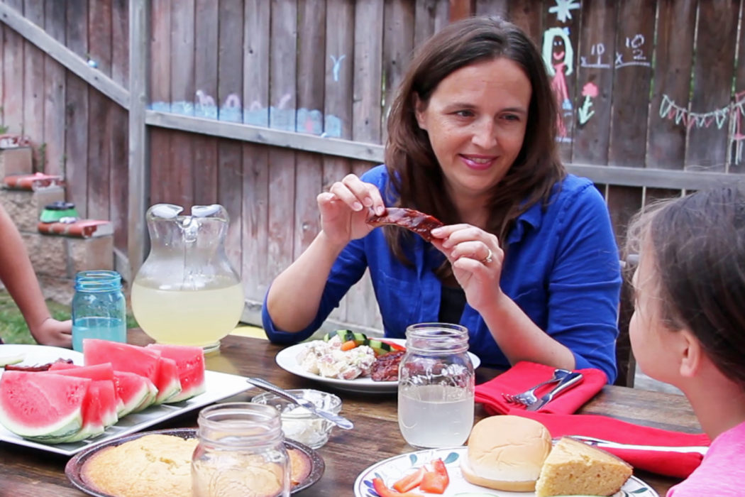 Jessica sitting at a table with a plate of food, holding a rib.