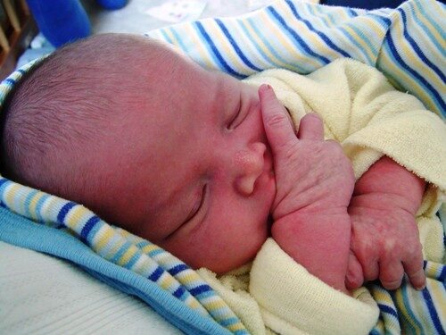 A baby lying on a striped blanket sucking his thumb.