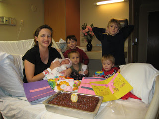 A mom with newborn sitting at a hospital bed with a birthday cake, and kids behind.