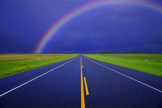 A rainbow in the background of a road with a yellow line down the center.