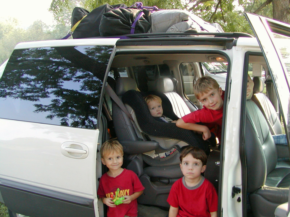 Three boys leaning out of a white minivan with baby in carseat.