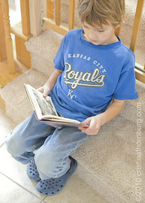 A little boy in a blue Royals shirt sitting on the bottom step, reading a book.