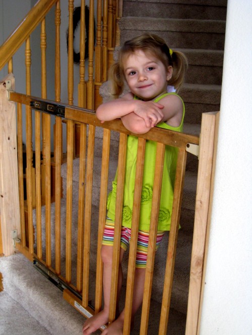 A little girl standing on baby gate on stairs.
