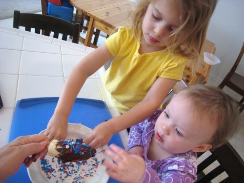 Little girls at kitchen counter dipping chocolate banana in sprinkles.