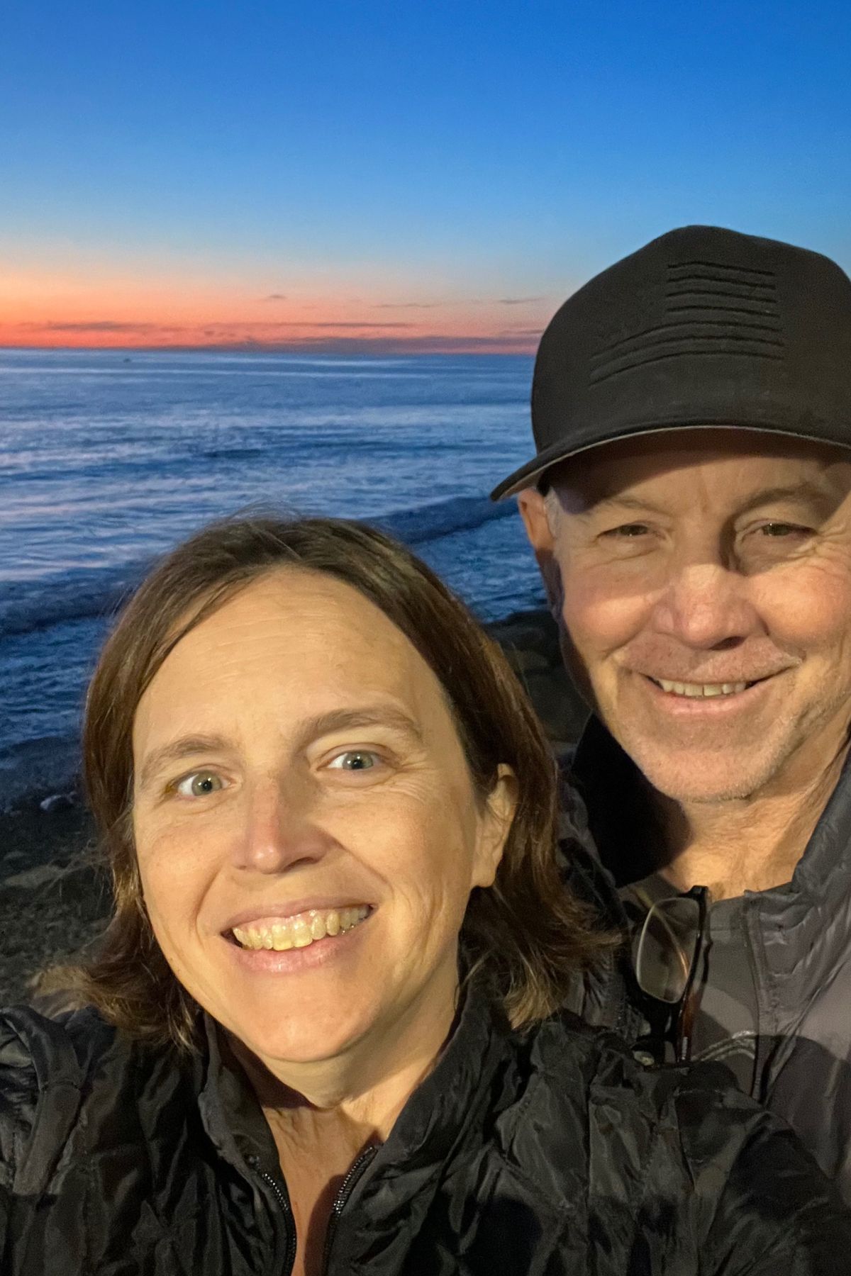 woman and man smiling at camera with ocean sunset in the background.