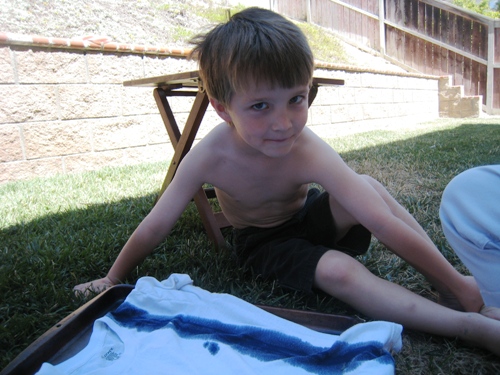 A little boy sitting on the grass with tie dye shirt on the ground near him.