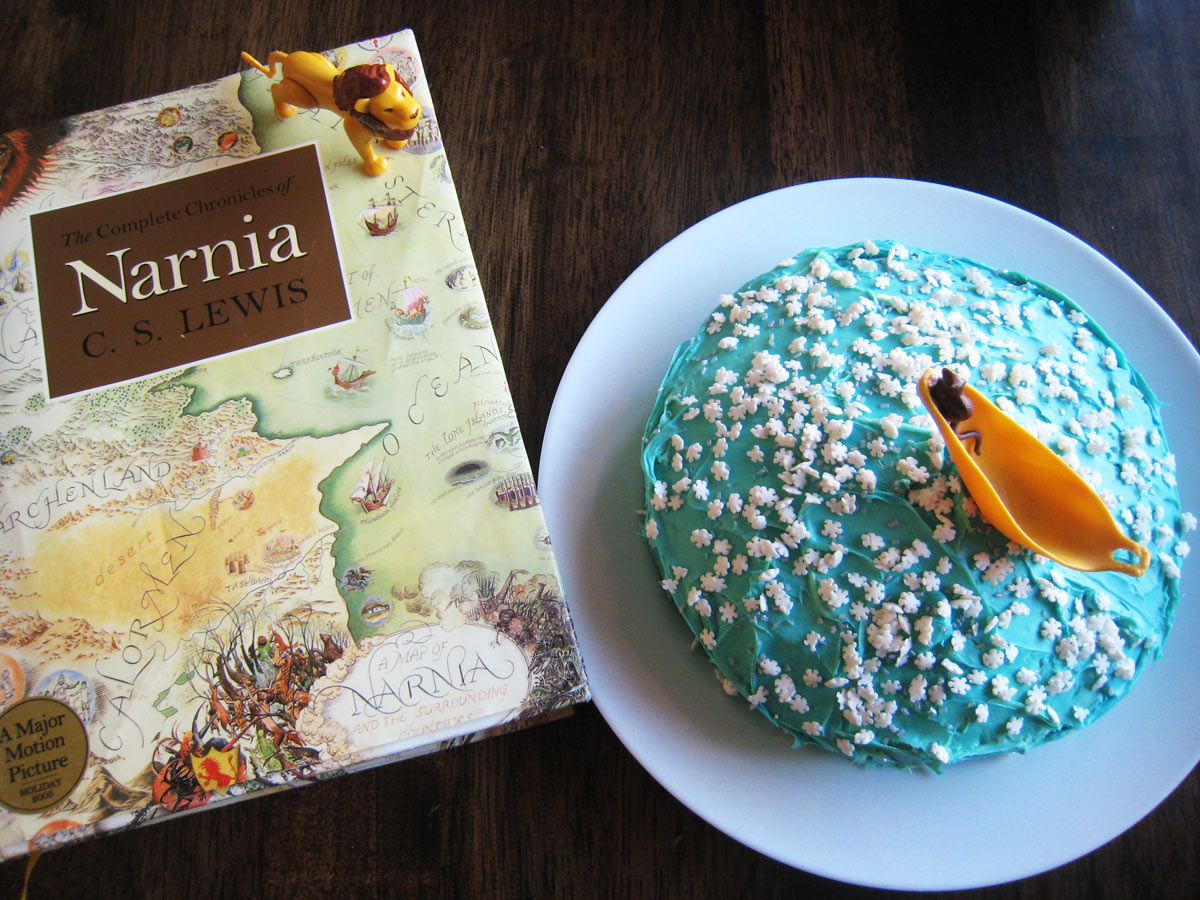overhead shot of narnia book with lion figure next to a cake on a platter with a mouse and boat atop it.