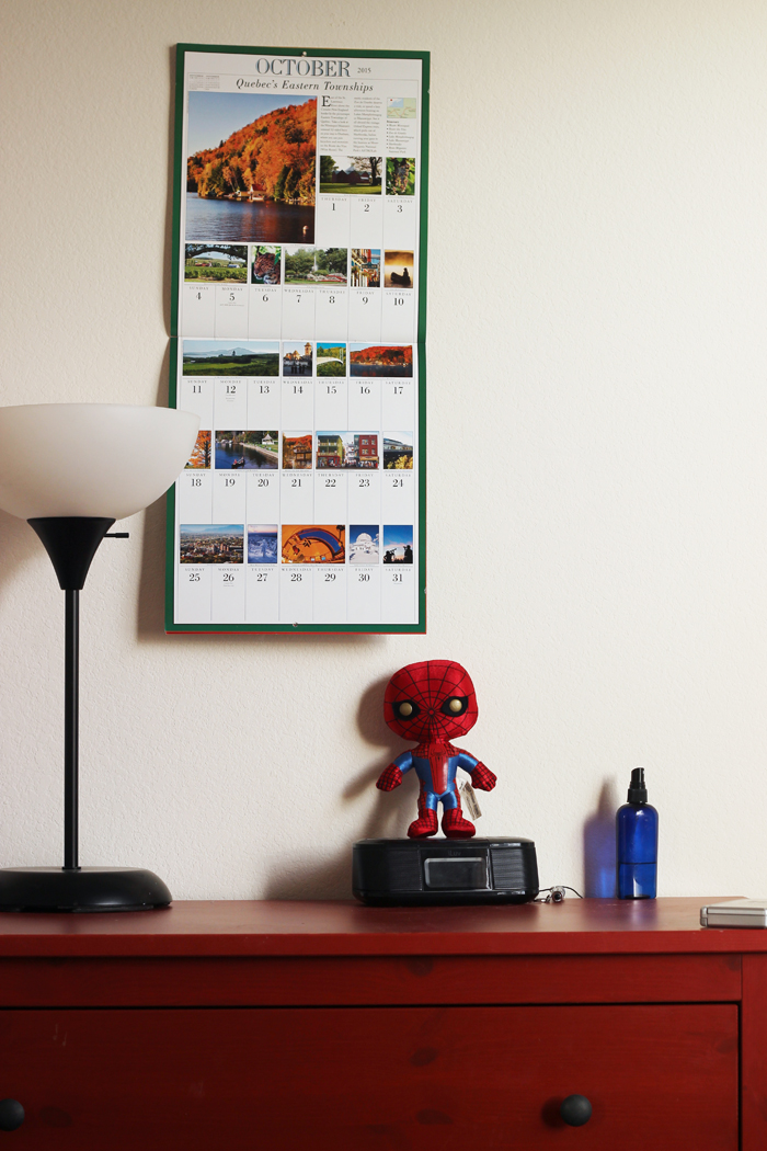 A dresser with a lamp, alarm clock, and spider man doll, with calendar on wall.