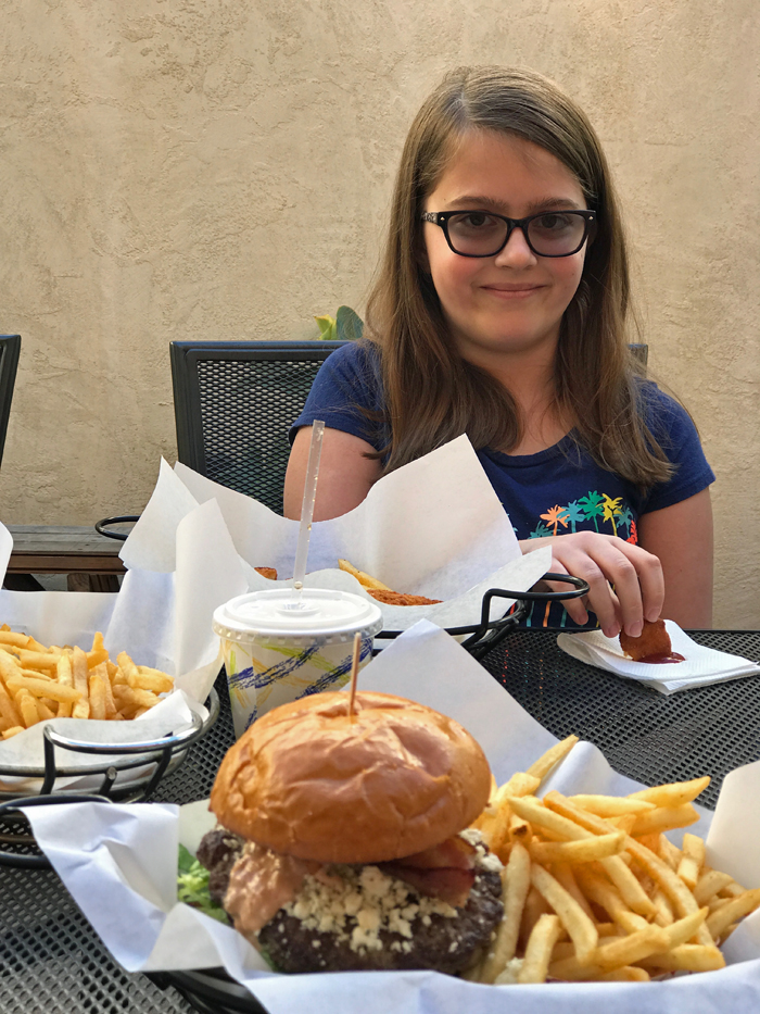 A girl sitting at a table with a plate of food, a burger in the foreground.