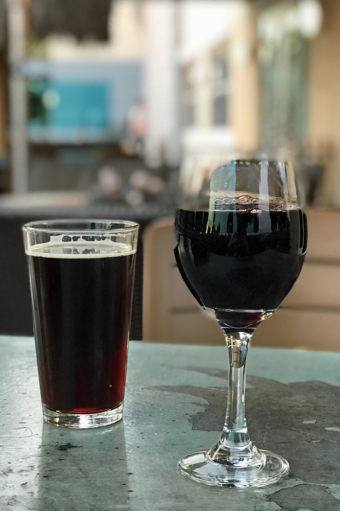 pint of beer next to glass of wine at a restaurant with fire pit in background.