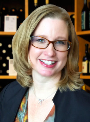 A woman in glasses looking at the camera, with wine in background.