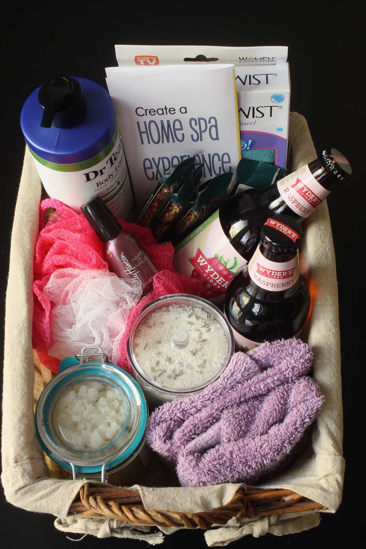 overhead shot of cloth lined gift basket filled with spa items, snacks, and hard cider as well as a copy of home spa experience how-to printable.