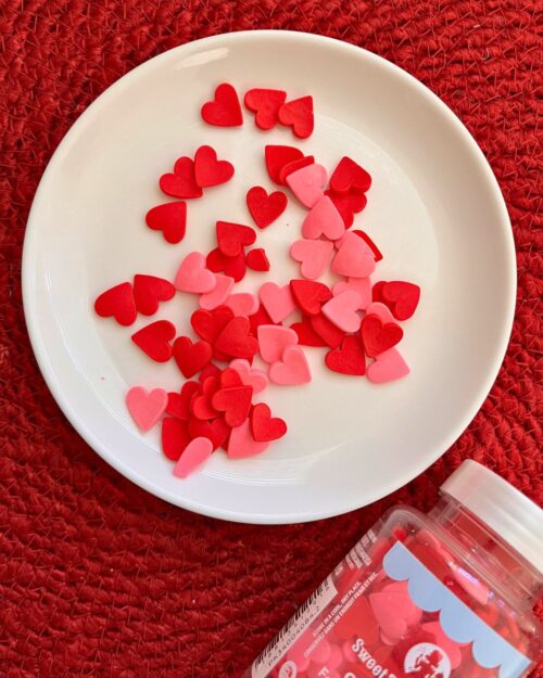 Pink and red heart sprinkles on a white plate on a red surface with the jar nearby.