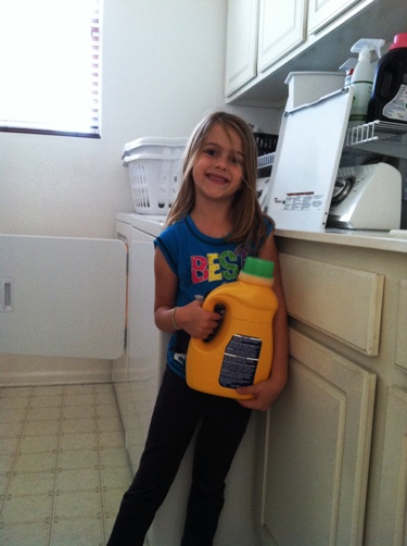 A girl holding a bottle of laundry soap leaning on a counter in the laundry room.