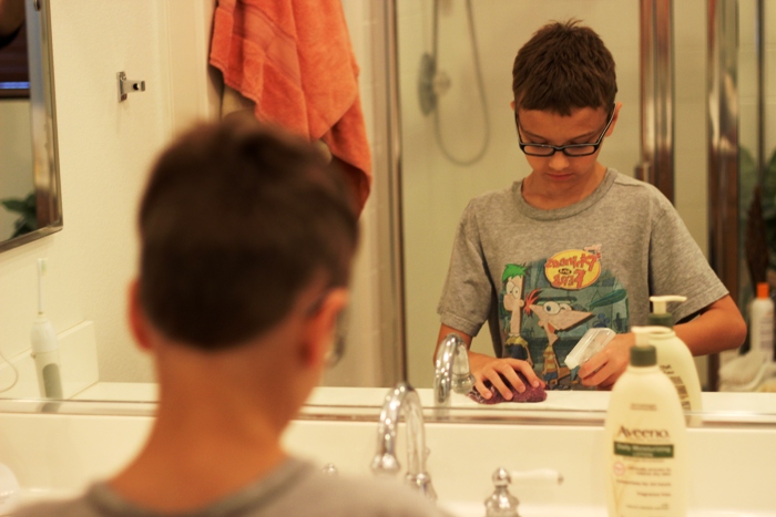 A boy cleaning the bathroom in front of a mirror.