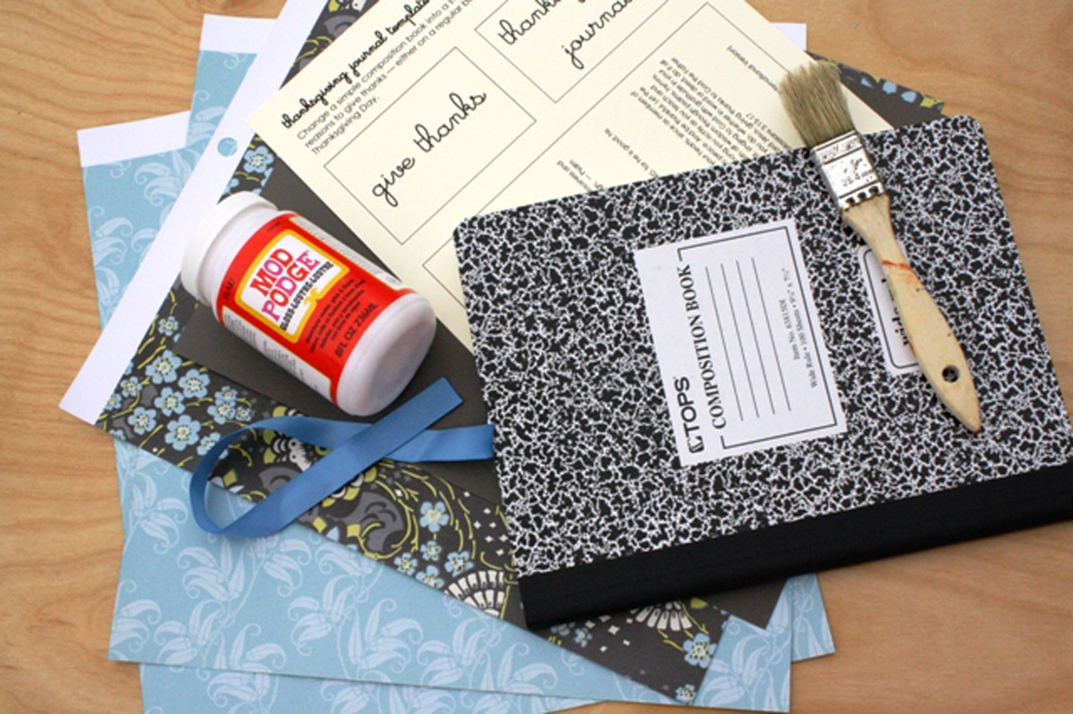 scrapbook papers, composition book, mod podge, printable labels, and paint brush laid out on the table top.