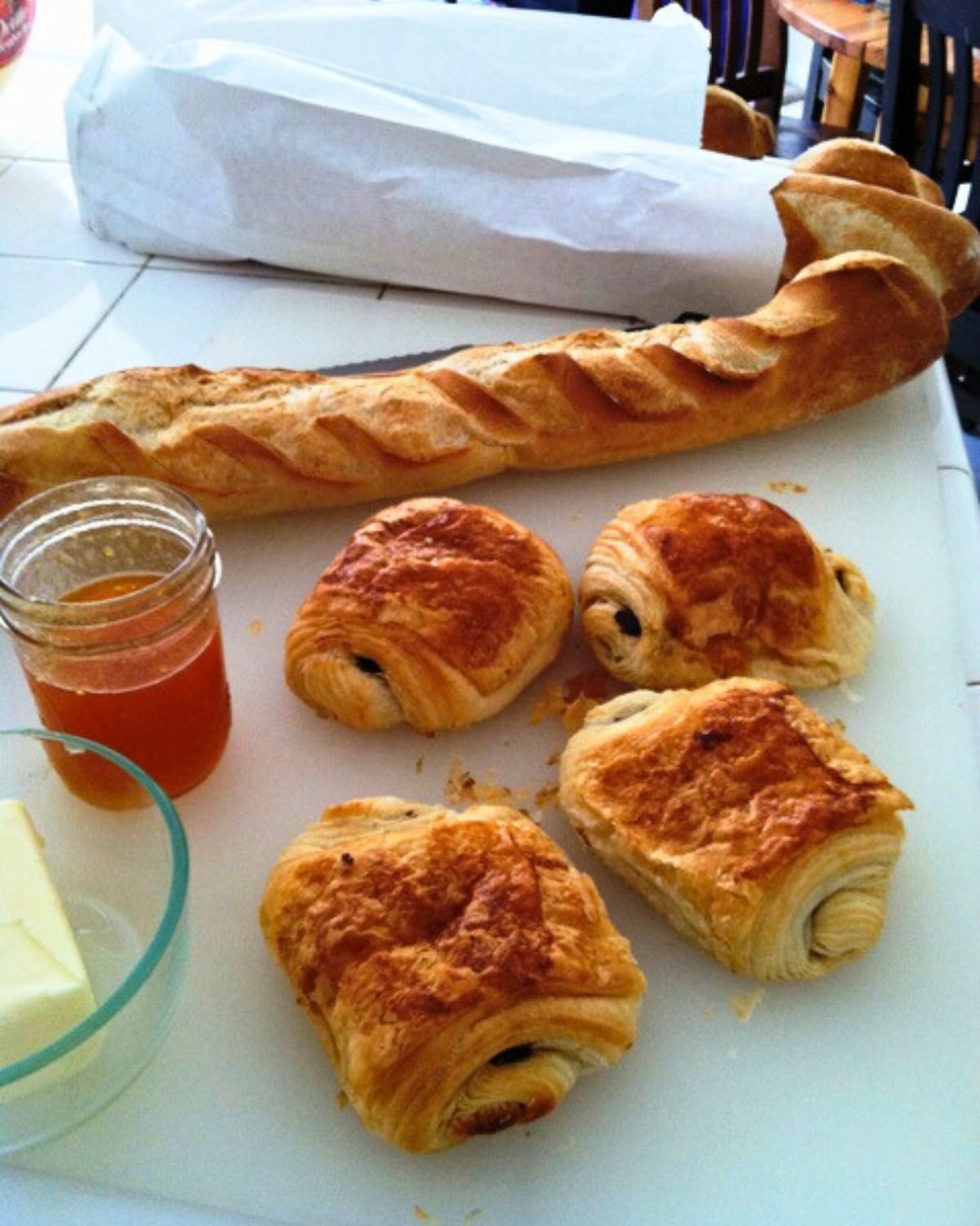 baguette and chocolate croissant laid out on white cutting board with bread and jam.