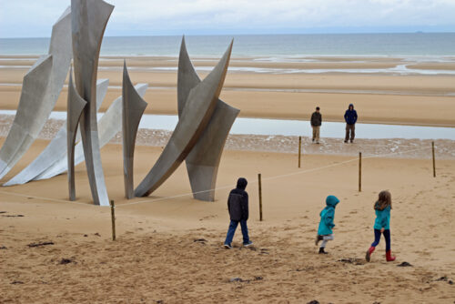 A group of kids standing on Omaha beach near a monument.