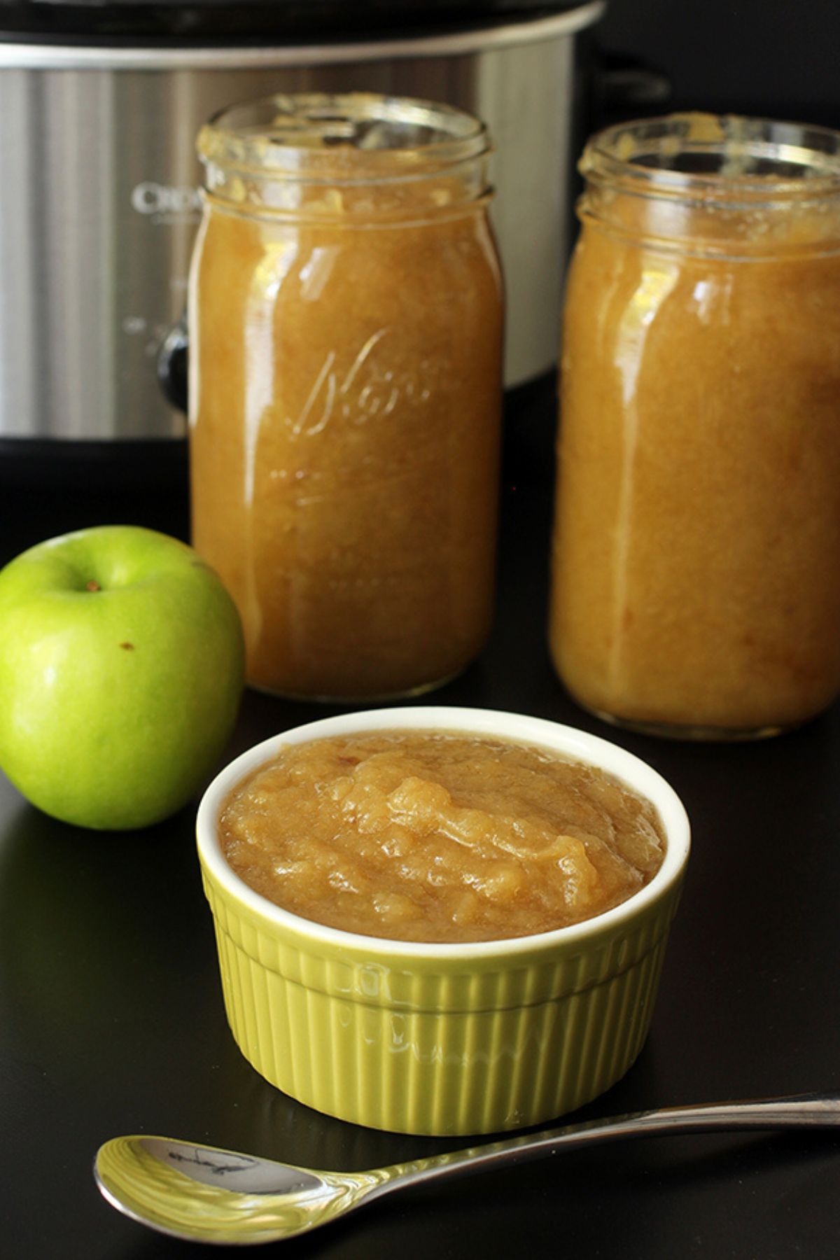 a dish of applesauce next to two large jars of applesauce with a green apple and a spoon.