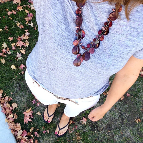 A woman wearing a necklace, tshirt, white jeans, and flipflops with leaves on ground.