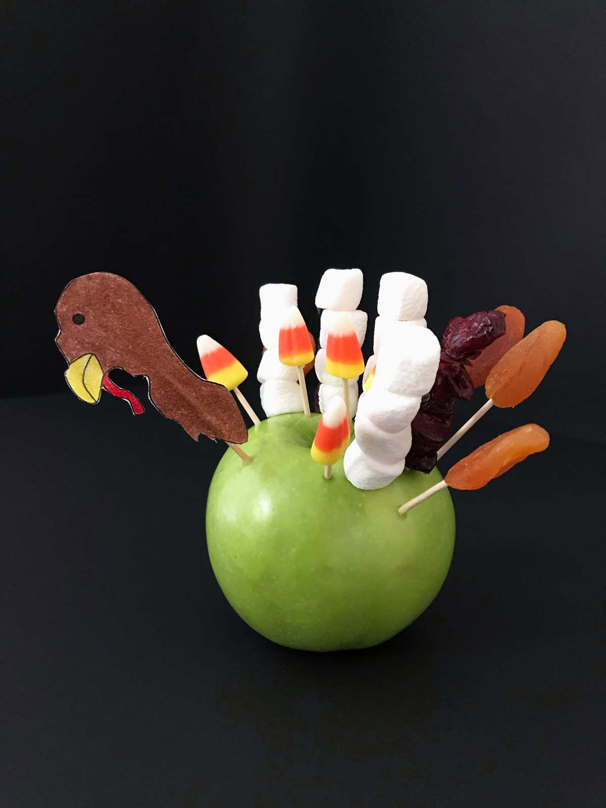 green apple decorated as a turkey with toothpicks, marshmallows, candy corn, and dried fruit.