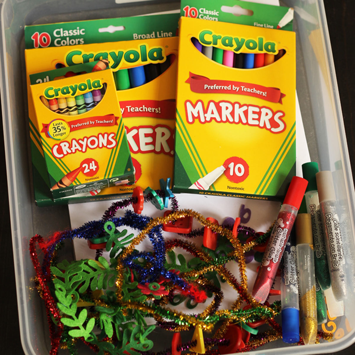 box of supplies including crayons, markers, glitter glue and pipe cleaners.