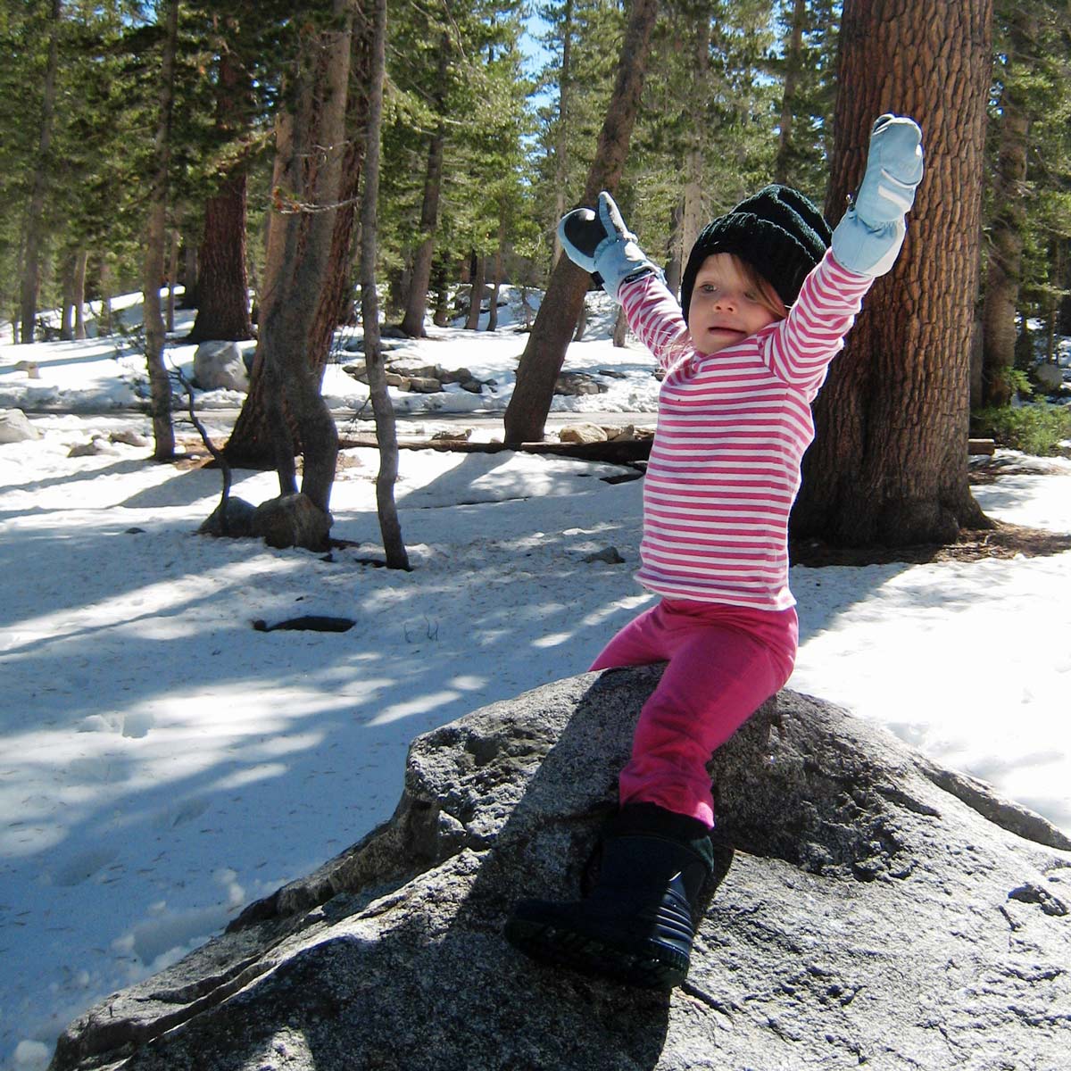 little girl with hat and gloves sitting on rock in a snowy forest area.