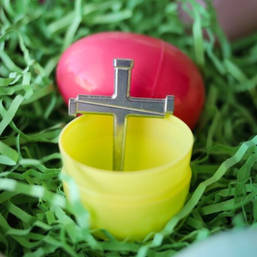 plastic yellow egg with a small metal cross made of nails sitting inside it, on grass with other plastic eggs nearby.