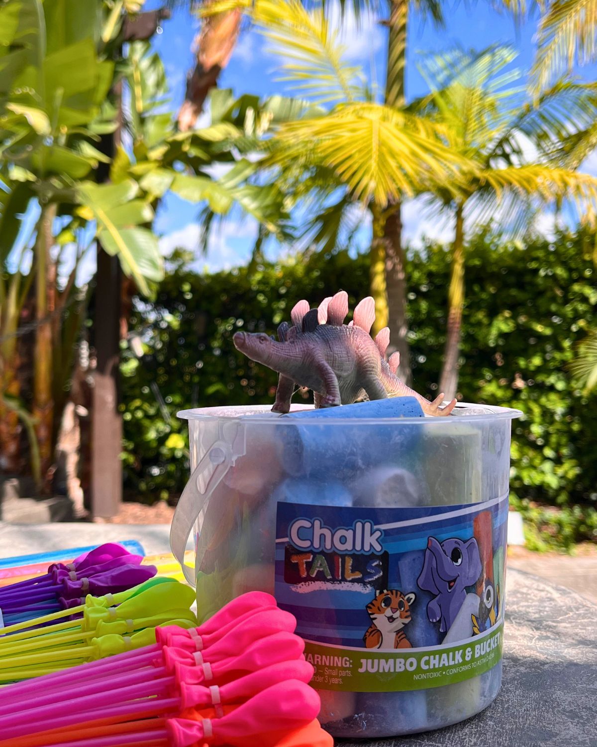 plastic dino atop bucket of sidewalk chalk by pile of balloon bunches.