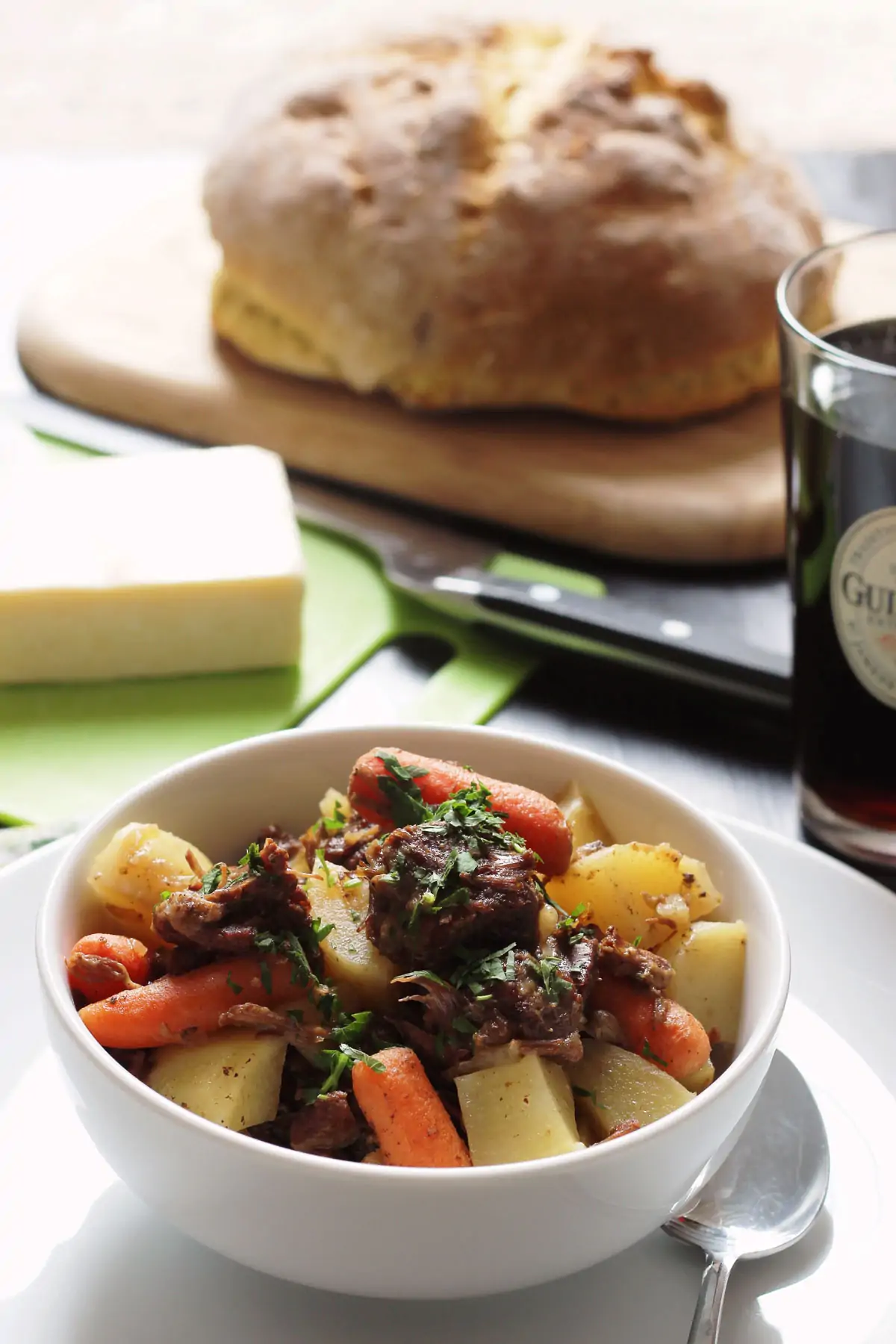 a bowl of irish stew with a loaf of soda bread and a glass of guinness in the background.