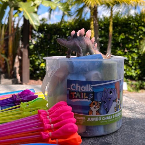 bunch of balloons next to bucket of sidewalk chalk with a toy dino on top.