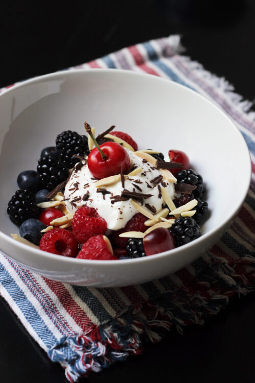 A bowl of fruit on a plate, with yogurt, chocolate, and nuts.
