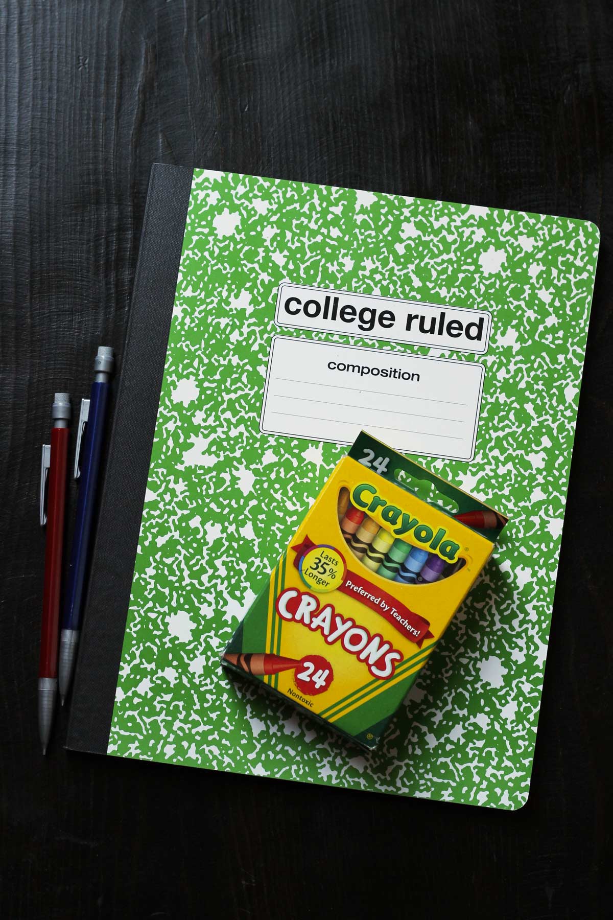 box of crayons atop green composition book with pencils nearby on black table.