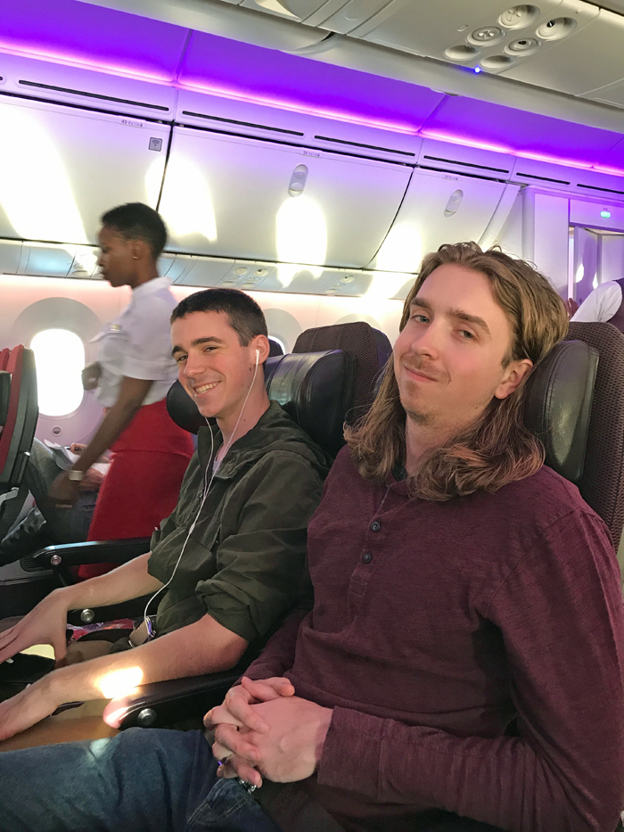 Two teen boys in their seats on a plane with the stewardess in the background.