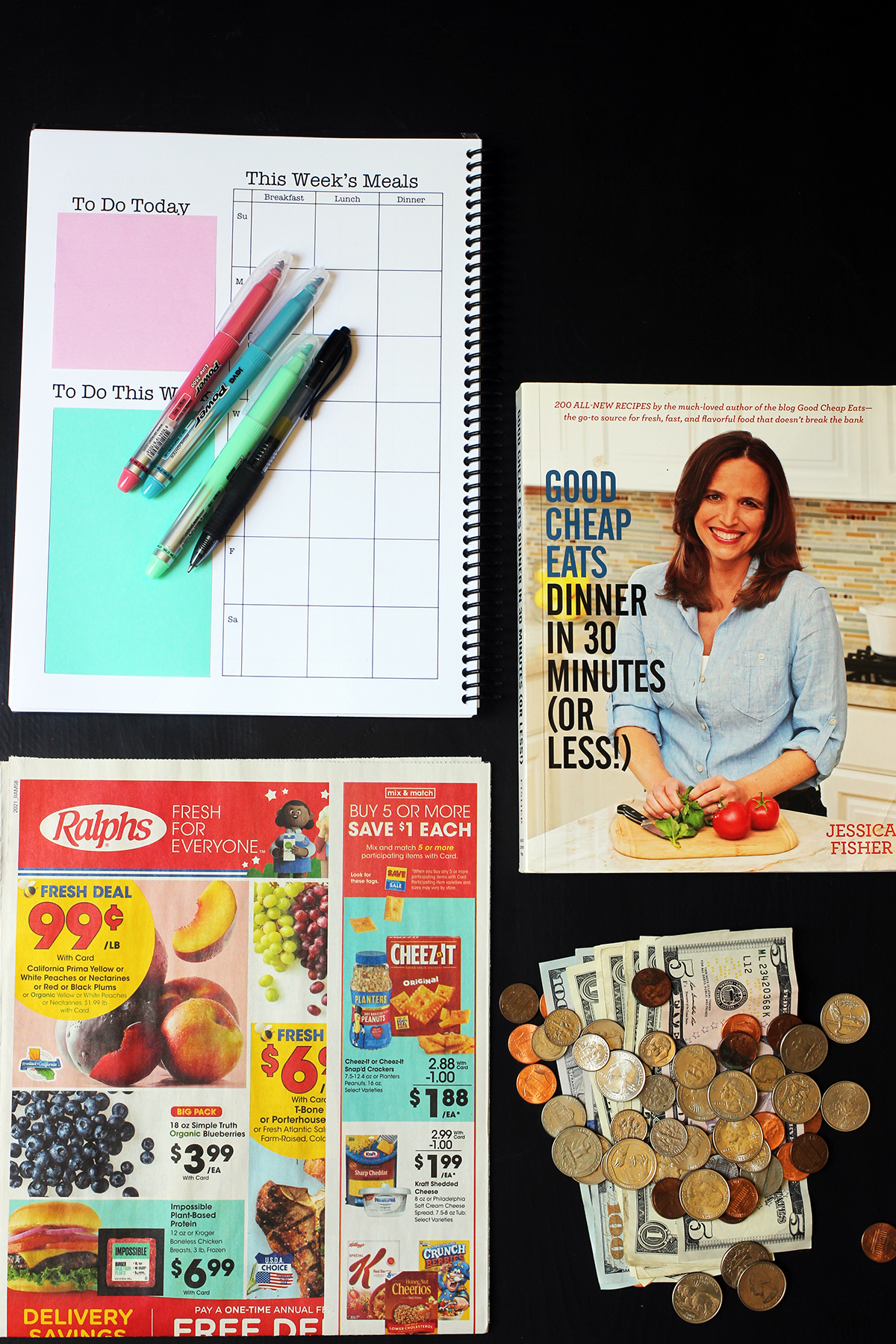 flatlay of planner, grocery ad, cookbook, and pile of money on black table.