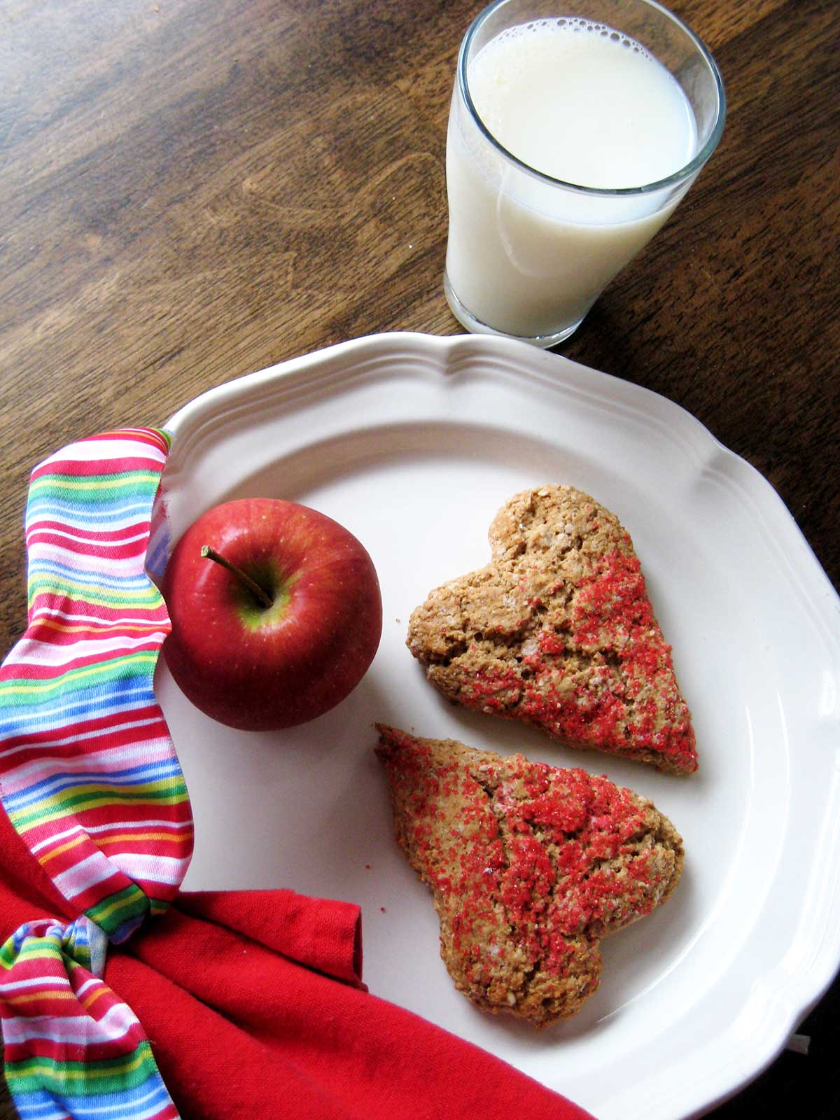 heart-shaped scones on a plate with an apple, the napkin wrapped in a bow, a glass of milk nearby.