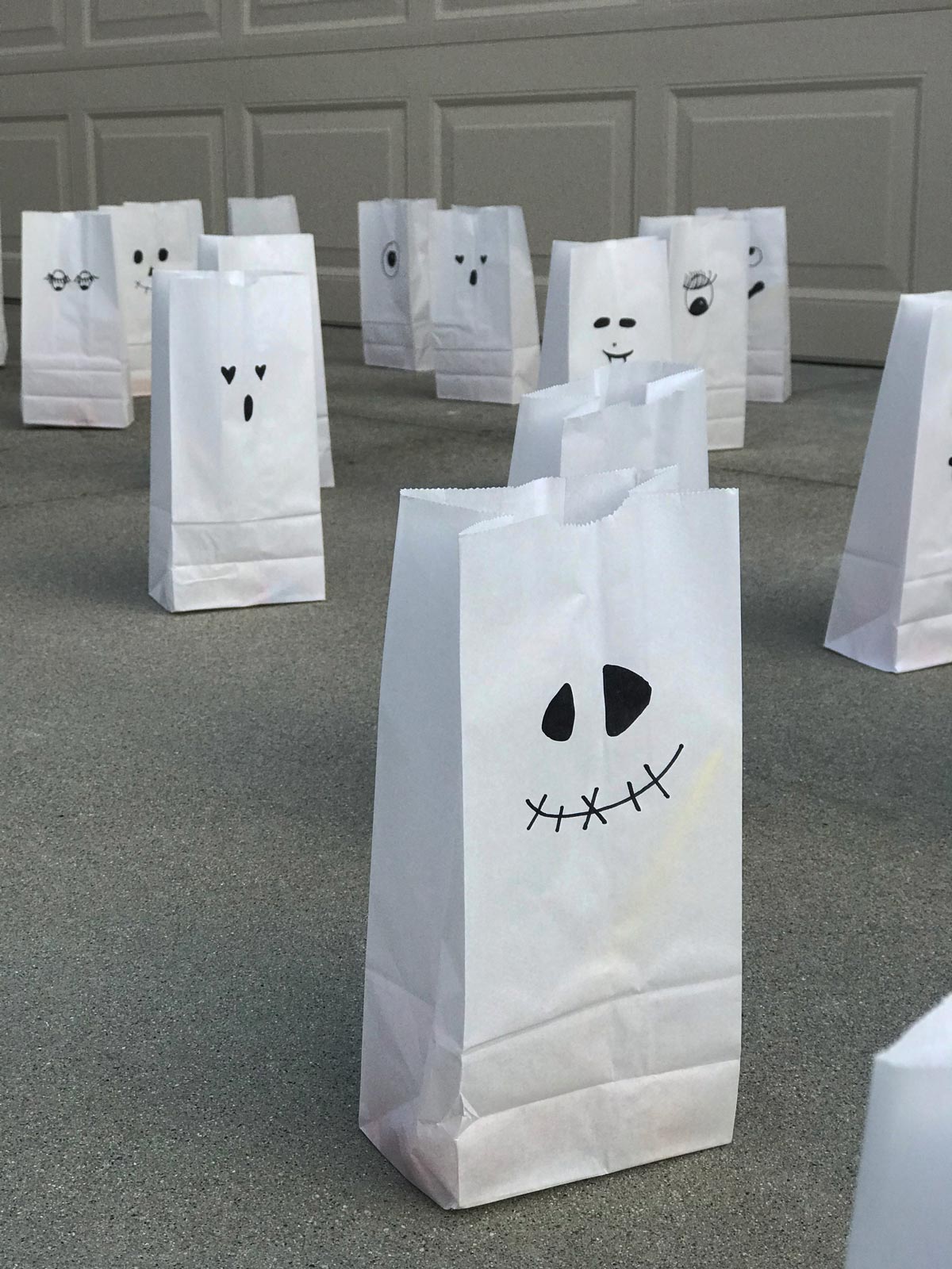 white paper bag ghosts set up on driveaway in an array, one looks like Jack Skeleton.