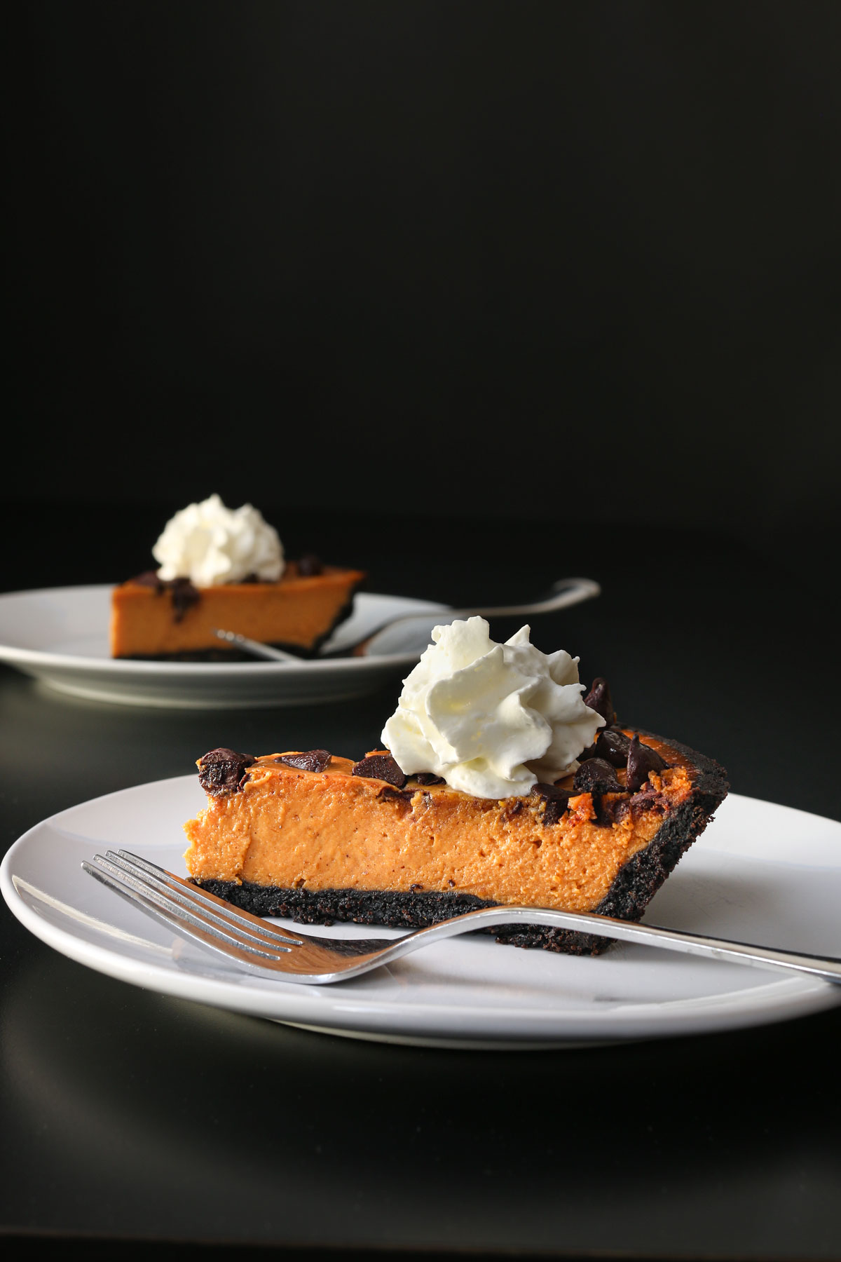 slices of pumpkin pie with chocolate crust and chocolate chips topped with fluffy whipped cream on plates with forks on a black table top with a black backdrop.