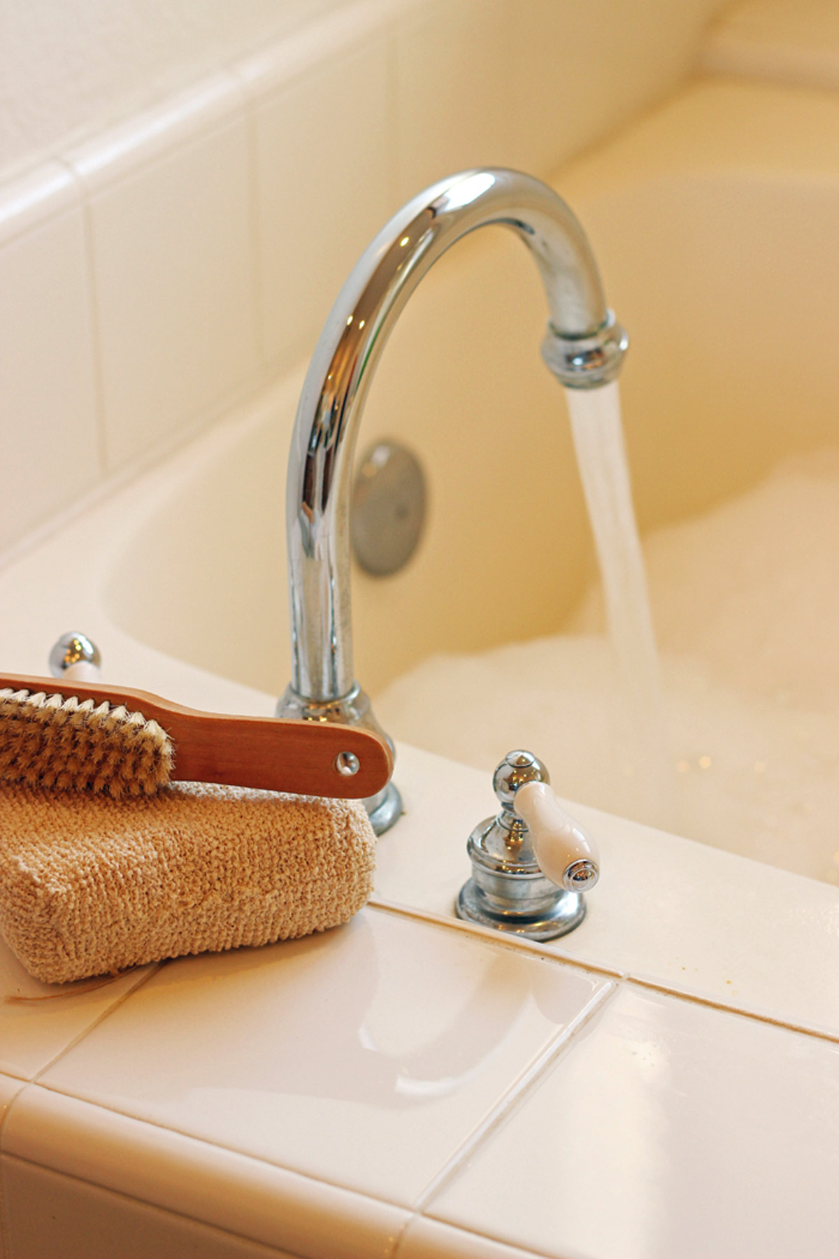 running water into a bubble bath in a white towel bathroom, there is a loofah and a scrub brush on the tile near the faucet, the sun is shining in.