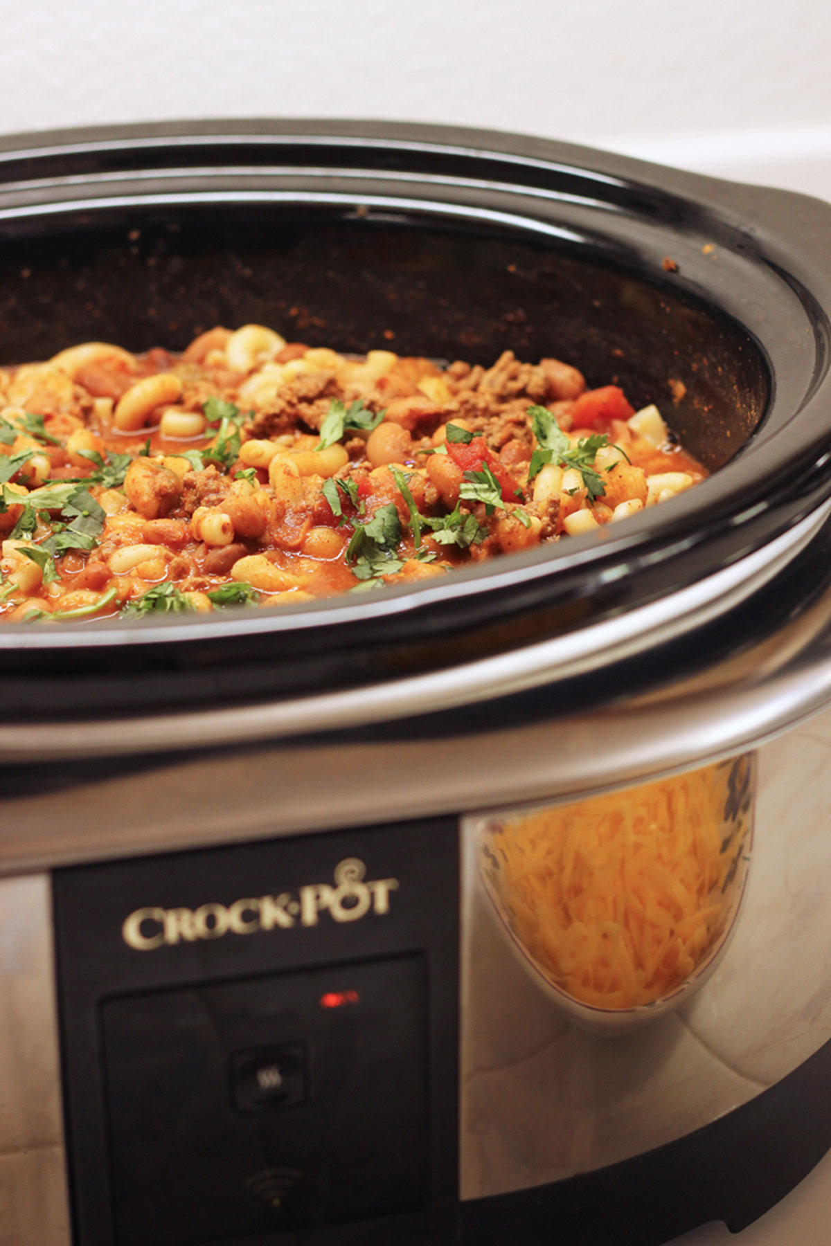 crockpot full of chili mac with bowl of shredded cheddar in its reflection.