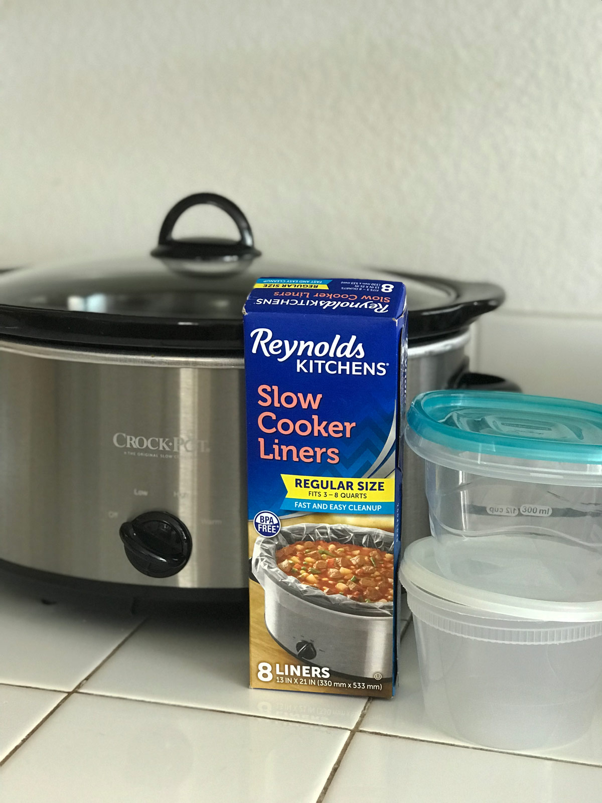 crockpot, box of liners, and plastic containers on white tile countertop.