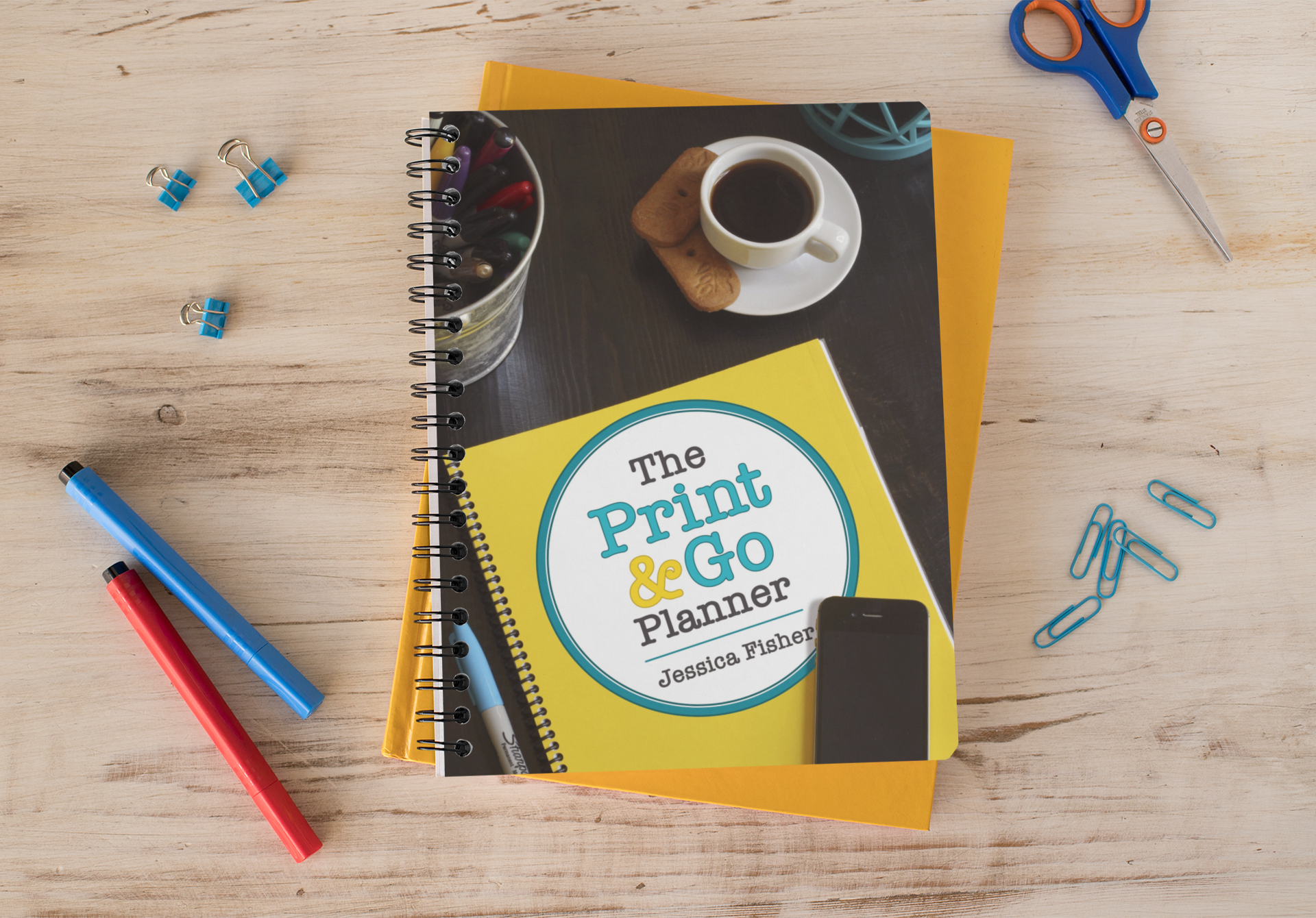 a mockup of print and go planner on wood table with markers, paper clips, and a pair of scissors.