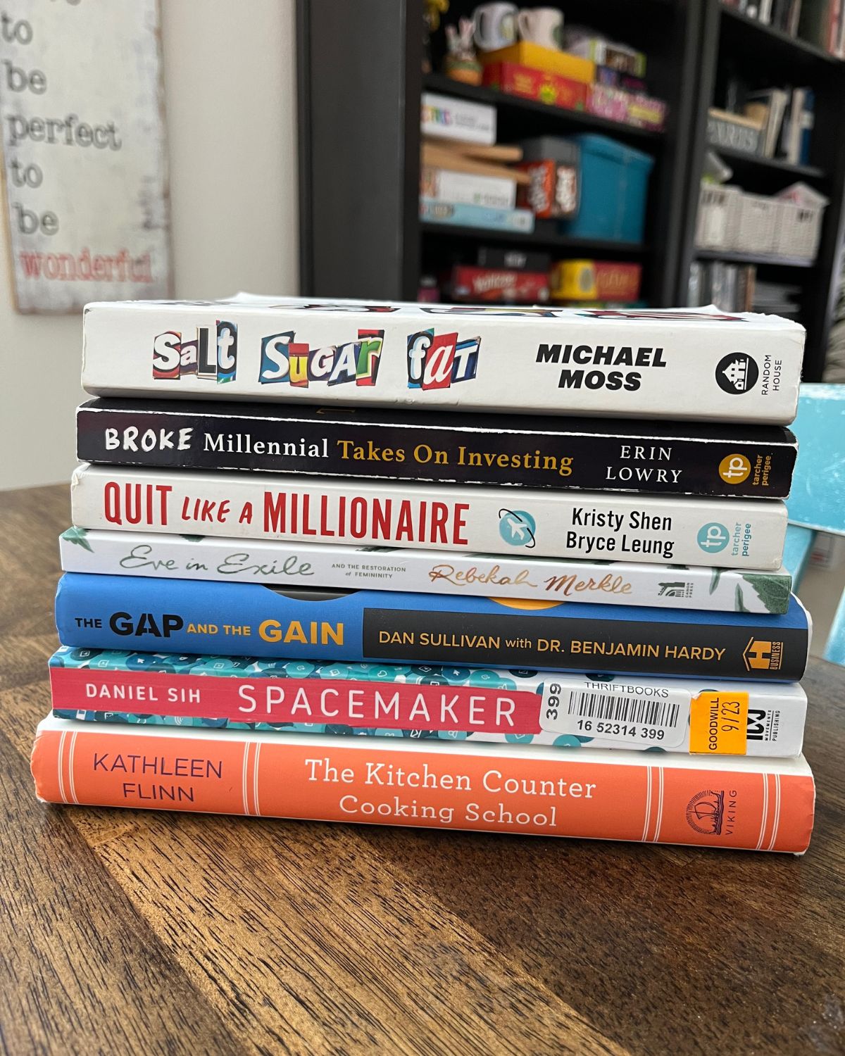 stack of books on a wood table with bookshelves in the background.