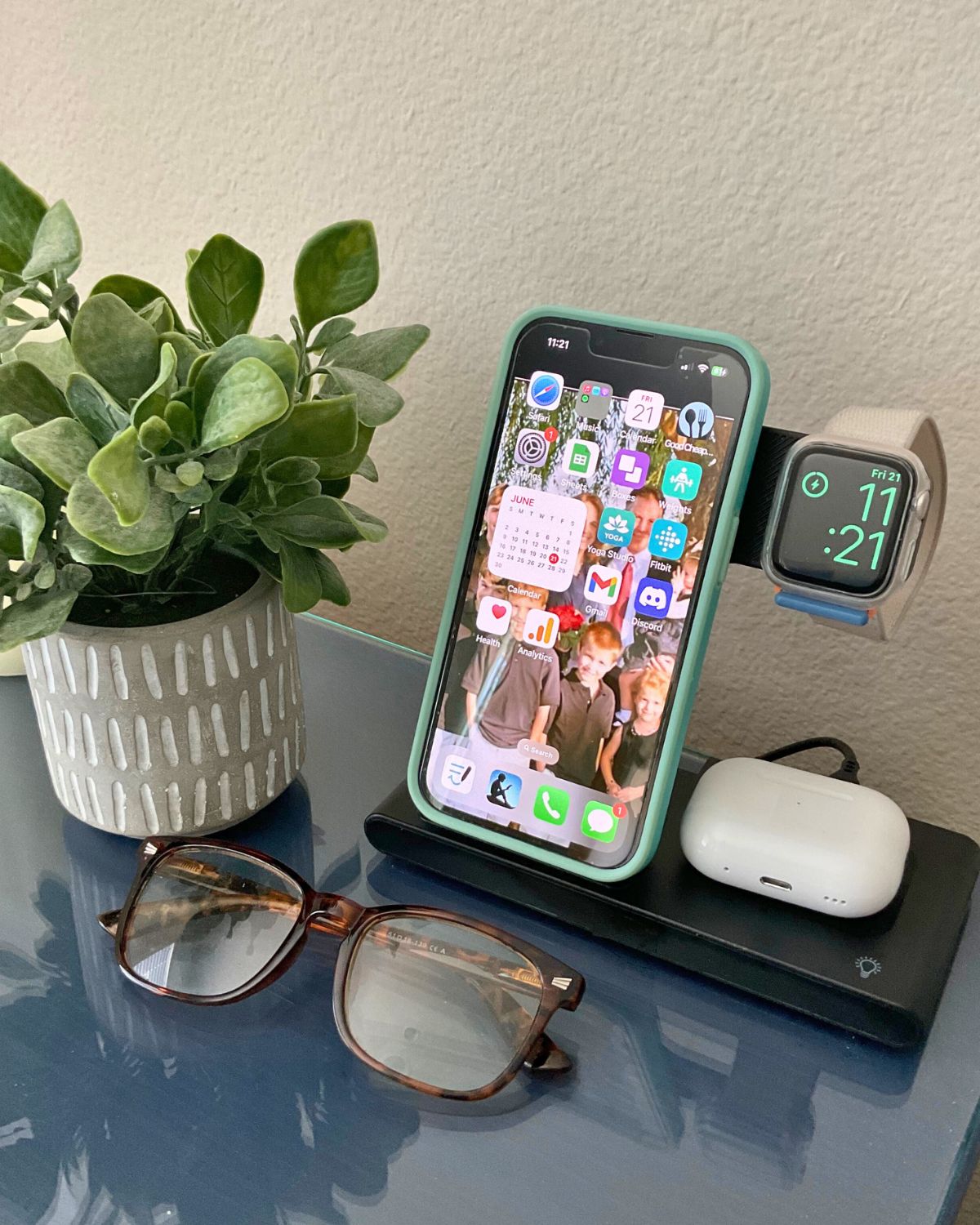 bedside phone charger with devices charging, glasses and plant nearby.