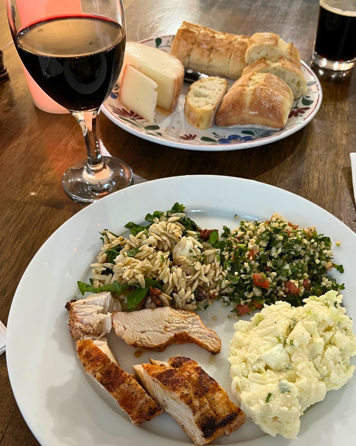 dinner plate with three salads and grilled chicken, cheese plate and wine glass nearby.