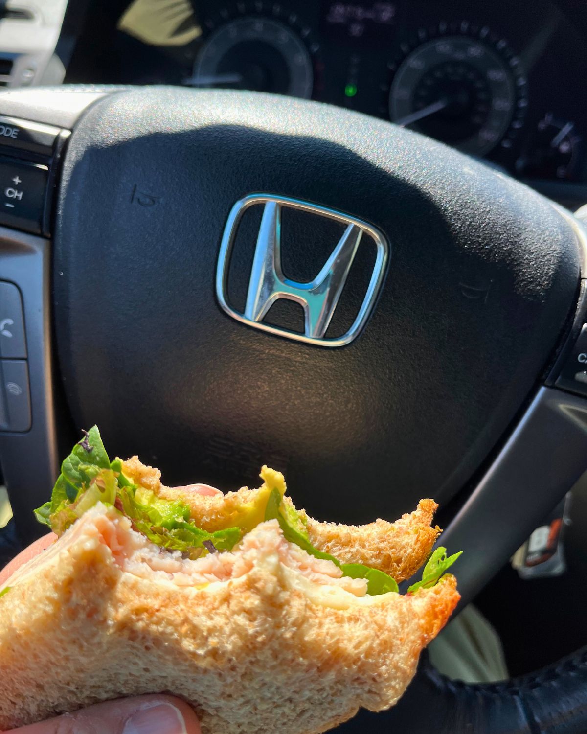 a half eaten sandwich in front of a honda steering wheel.