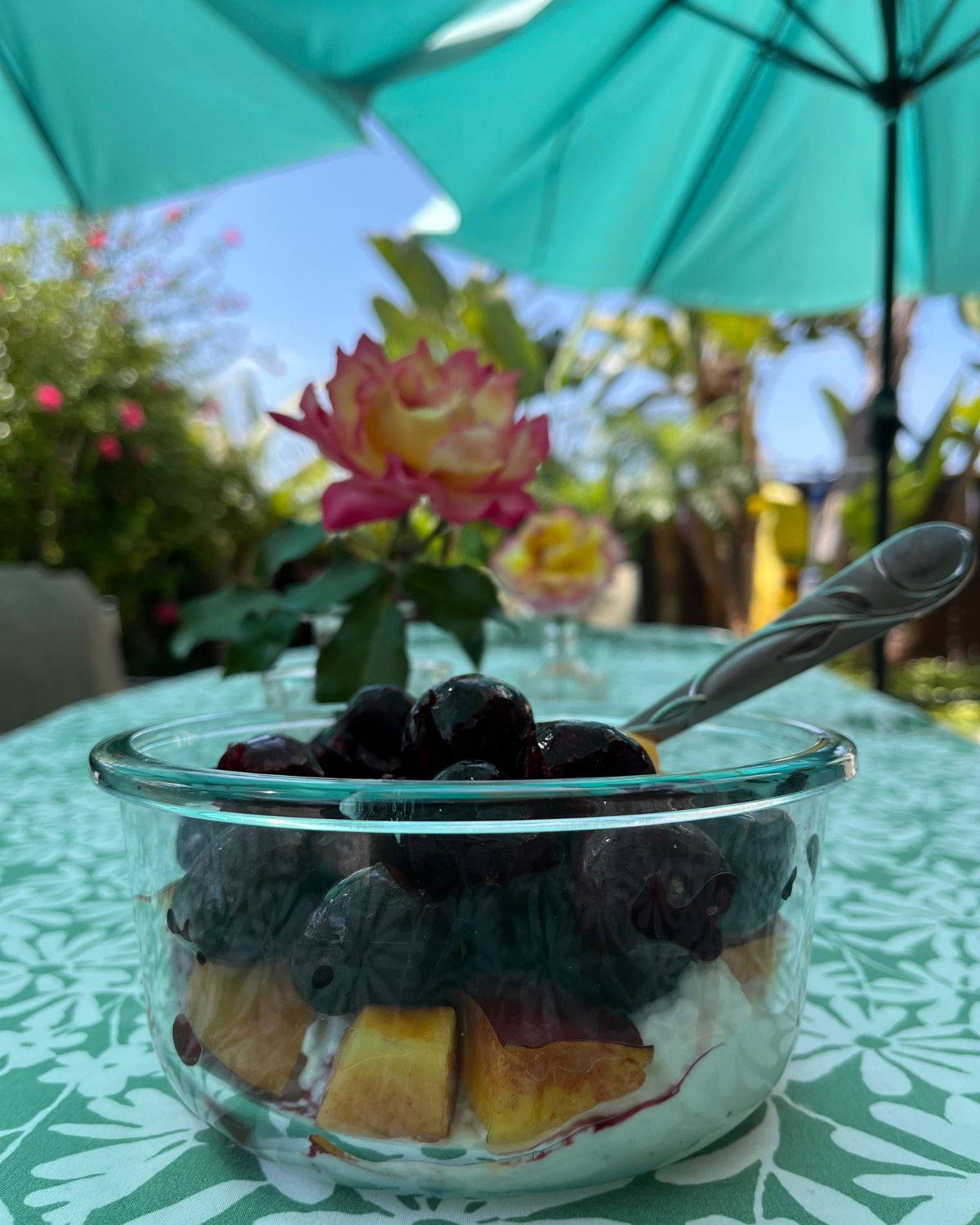bowl of fruit salad on table with rose bud vase and sun umbrella in background.