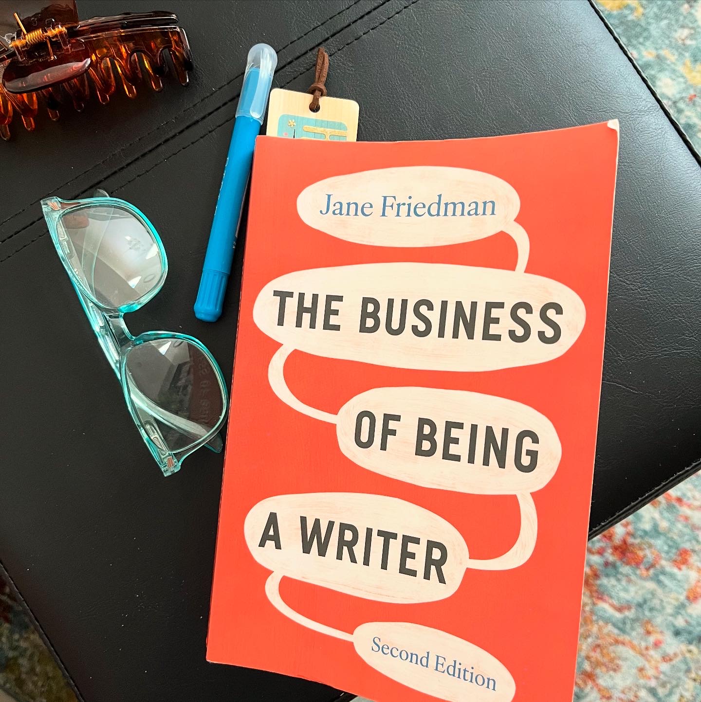 paperback copy of The Business of Being a Writer on ottoman with reading glasses and highlighter.