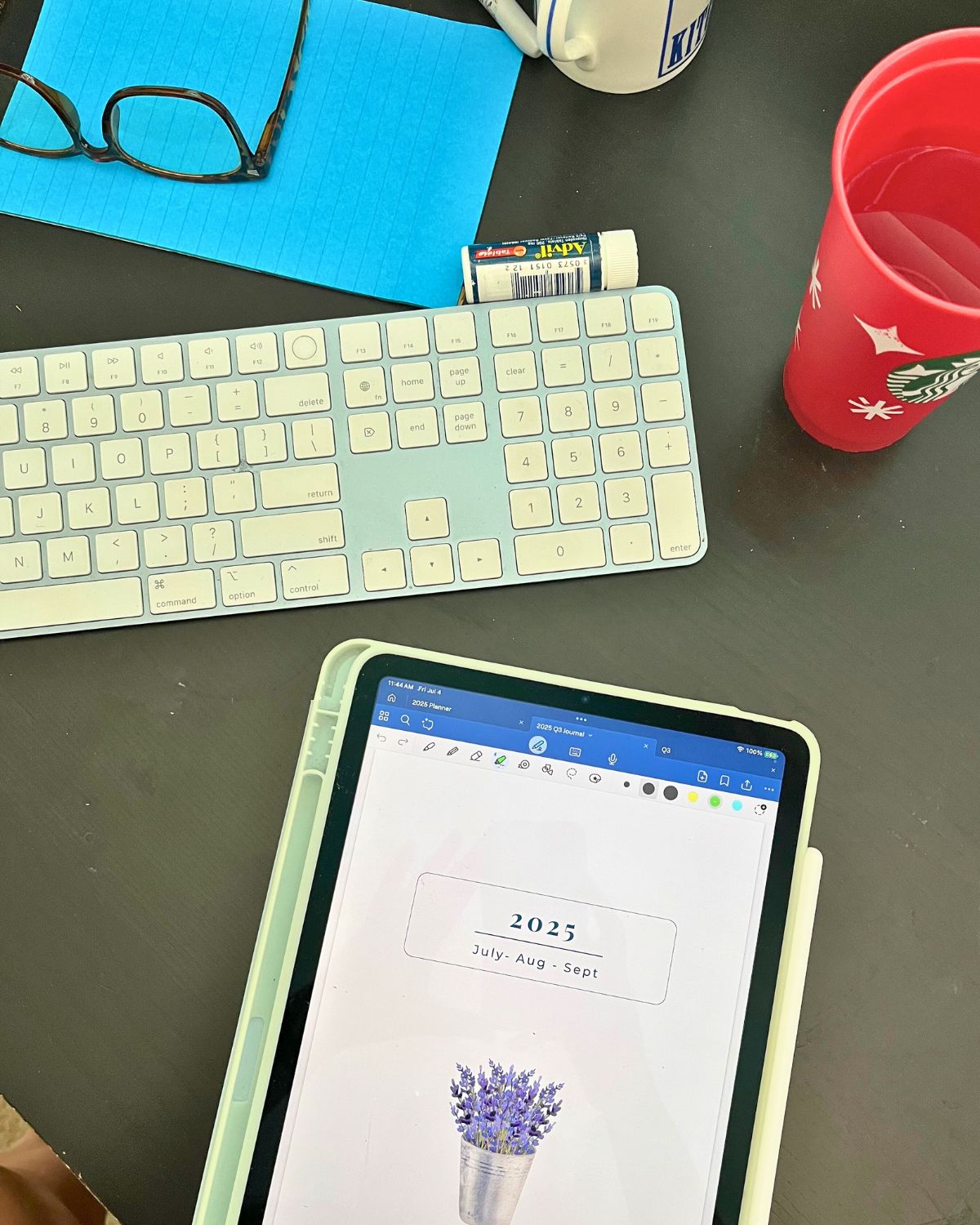 desktop with notepad, glasses, advil, keyboard, water glass, and iPad.