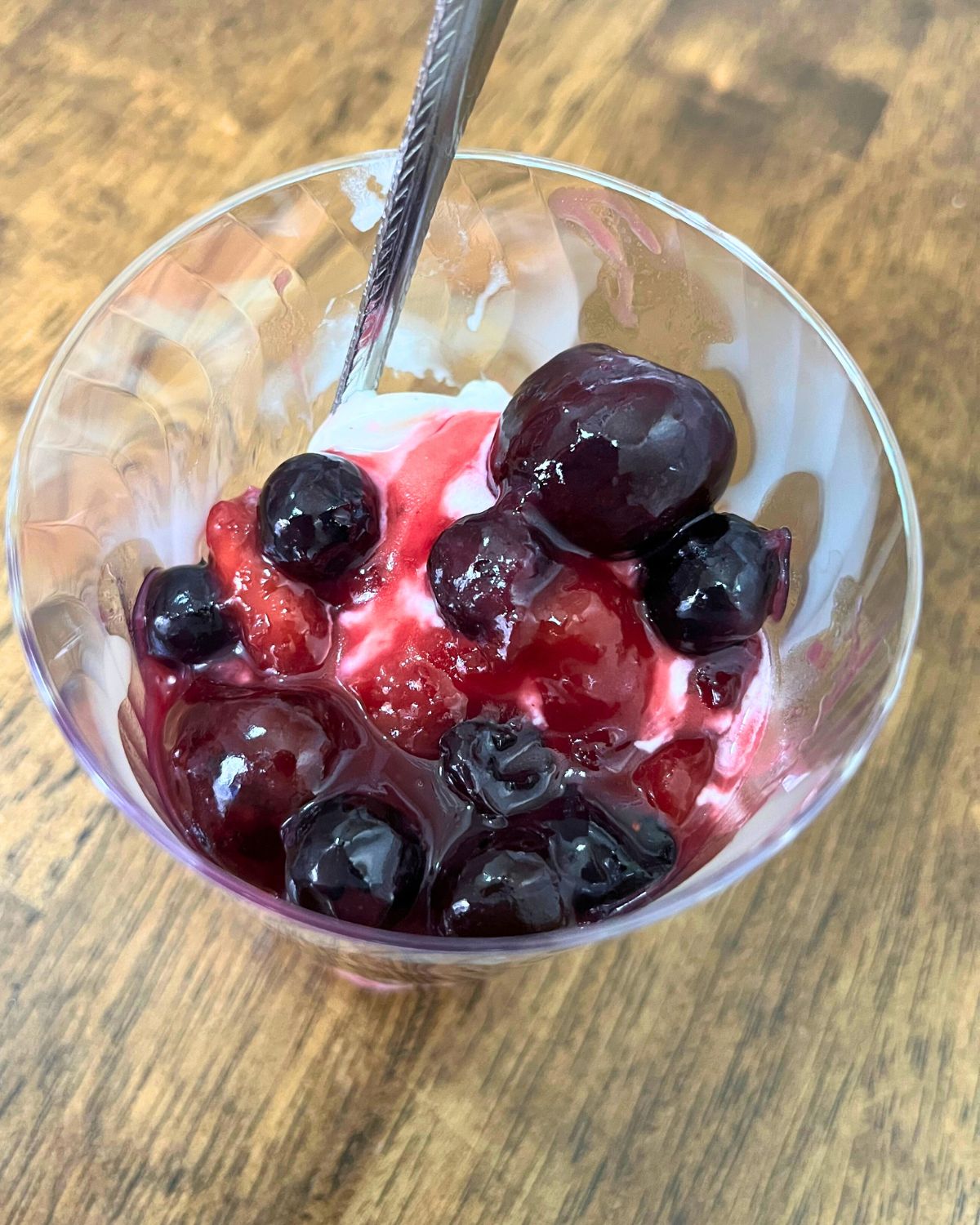 A plastic cup of ice cream and fruit lumpy with a spoon on a wooden table.